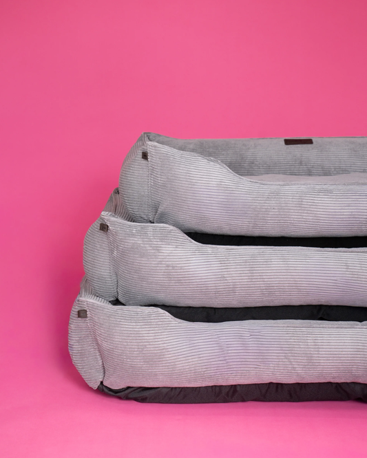 Three stacked grey corduroy memory foam bolster pet beds on a vibrant pink background.