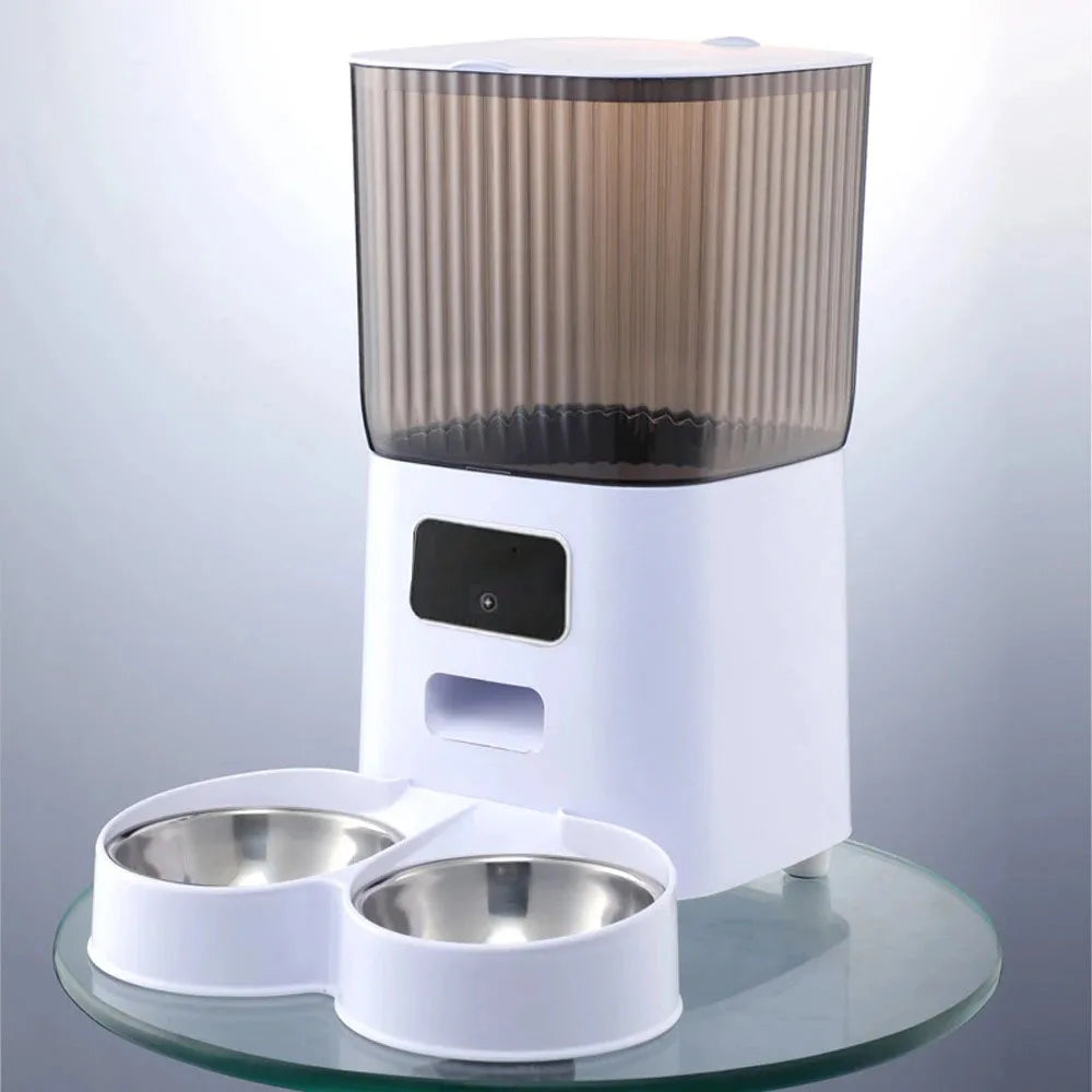 White 5L smart cat feeder with smoky ribbed hopper, dual stainless steel bowls, and a built-in camera on glass table.