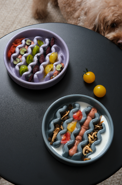 Purple and blue-grey ceramic macaroni slow feeder bowls filled with food on a black table with a dog nearby.